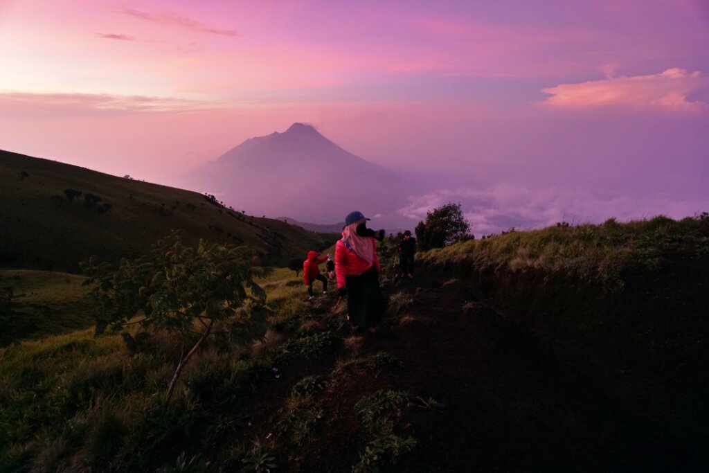 Perjuangan para pendaki meniti tanjakan di tengah sabana menuju puncak Gunung Merbabu, dengan latar belakang Gunung Merapi